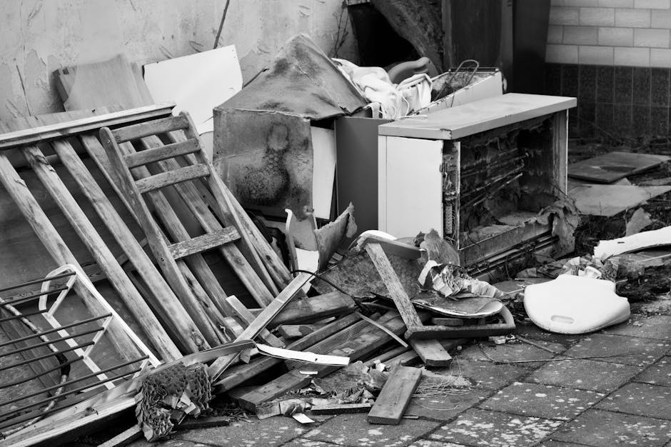 The black and white image depicts a cluttered outdoor scene featuring a pile of discarded household items and rubbish on a paved surface. Prominently, there are two wooden garden chairs with slatted backs and seats, leaning against a wall on the left side of the image. Behind and around them, various debris includes broken wooden planks, a damaged metal-frame item, and pieces of torn cardboard or paper. To the right of the chairs, a partially open white cabinet or appliance housing debris is visible, with a large, discoloured metal object resting partially inside, showing rust and grime. There also appears to be a bulky plastic object, possibly a damaged container, situated on the ground towards the right side. The background includes a brick wall and a small section of a darker wall or fence. This scene is characteristic of an area awaiting rubbish removal or private waste collection services like those offered by Waste Clearance Streatham, illustrating the type of clutter that requires professional clearance solutions. Overall, the image captures a messy, neglected space with a variety of materials in different textures, such as wood, metal, and plastic, in a condition suitable for a rubbish removal service specializing in on-site clearance or alternative waste handling.