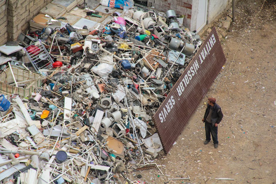 A person standing next to a large pile of mixed household waste and discarded items on an outdoor dirt surface, with numerous broken appliances, metal and plastic objects, and miscellaneous debris visible within the heap. The waste includes damaged electrical components, metal frames, plastic containers, and various unrecognisable household items scattered across the heap. The scene is surrounded by a brick wall on one side and a weathered wooden fence with a sign behind the waste that reads 'EXIT IN USE' in large white letters. The environment appears to be an outdoor storage or waste disposal site with uneven ground and no vegetation in sight. Natural daylight illuminates the scene, highlighting the messy and cluttered state of the rubbish, which is indicative of an independent waste clearance effort, potentially managed by Waste Clearance Streatham, as part of private increased rubbish removal services beyond standard municipal collection. The overall atmosphere is utilitarian, with the focus on discarded household waste and debris awaiting disposal or recycling.