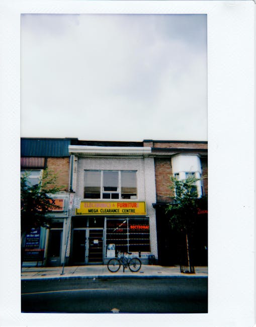 A two-storey commercial building featuring a large yellow sign reading 'Electronics & Furniture Mega Clearance Centre' mounted above the entrance. The facade includes a combination of brickwork and a white cladding panel beneath the window on the upper level. Large glass doors and windows form the storefront on the ground floor, with a black bicycle parked directly in front, leaning against a short metal railing. The sidewalk in front is clean, and the street has a smooth asphalt surface with a curb. Flanking the building on both sides are small trees, and to the right, part of another shop's sign is visible. The sky above is overcast, creating soft, diffused lighting. The image appears to be taken from a slightly low angle, capturing the entire facade and street scene. This setting connects to private waste handling services such as rubbish removal or clearance, as indicated by the clear presence of waste management opportunities in commercial areas, exemplified by Waste Clearance Streatham's professional services for clearing unwanted items from such properties.
