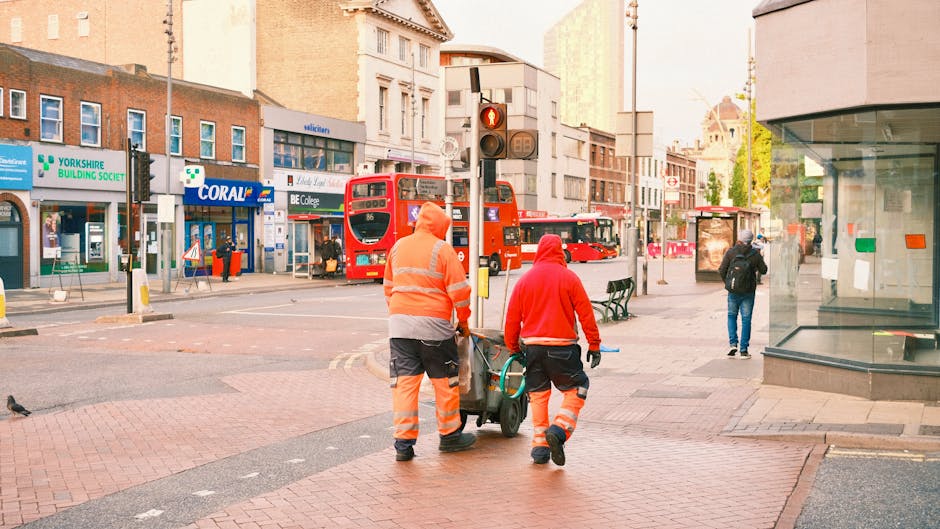 Two waste collection workers are walking along a city street during daylight, dressed in bright orange high-visibility uniforms with reflective stripes. They are pushing a small, wheeled bin or cart, which appears to be used for collecting waste or recyclables. The street background includes a row of buildings with commercial shops, such as a healthcare-related store with a green cross sign, a Coral betting shop, and other storefronts, some with signs visible. A red double-decker bus is stopped at a traffic light ahead, with pedestrians walking nearby, including a person in blue jeans and a dark jacket standing on the pavement, while another individual is visible further down the street. The environment is urban, busy, and well-lit, with a mix of modern and older architectural elements, and the overall scene suggests a typical street scene involving waste collection or street cleaning activities, aligning with private or alternative rubbish removal services provided by companies like Waste Clearance Streatham.