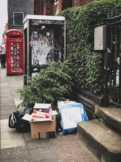 On a city sidewalk, a small pile of waste materials is placed in front of a black glass notice board and partially obscured by an untrimmed green shrub. The waste includes a black plastic bag, a cardboard box partially open revealing some contents inside, and a large blue and white cardboard box with printed symbols and text, indicating packaging or home delivery items. Behind the collection, there is a traditional red phone box and a black metal fence with dense green foliage growing along it. The scene is set on a paved pavement with a set of concrete steps leading up on the right side, suggesting this is an urban area with residential or commercial buildings nearby. The lighting appears natural, indicating daytime with a neutral, unobtrusive atmosphere, reflecting typical private waste disposal in an urban environment. The presence of waste outside a notice board and near the hedge suggests an alternative collection or rubbish pickup service facilitated by Waste Clearance Streatham, catering to private or independent rubbish removal needs.