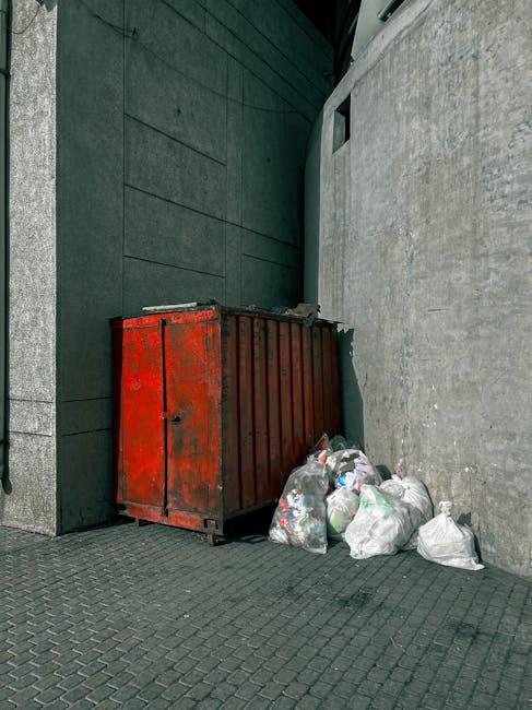 A large, rectangular, metal waste skip painted red with visible rust patches and chipped paint, positioned against a dark green textured wall on the left and a light grey concrete wall on the right. The skip is situated outdoors on a paving surface composed of small, dark grey interlocking bricks. In front of the skip, there are several white plastic garbage bags filled with household waste, some of which are tied at the top, while others are loosely closed, with a few secured with ties or twisted plastic. The bags and the skip appear to contain general rubbish, including possibly paper, plastic, and household discards. The background includes a small, narrow, rectangular window or vent above the skip on the concrete wall. The scene is lit naturally, with subtle shadows indicating daylight. This visual depiction suggests an instance of private waste collection or rubbish removal, possibly facilitated by a local waste management service such as Waste Clearance Streatham, known for handling on-site waste disposal in urban environments, integrating with alternative rubbish pickup options outside standard municipal services.